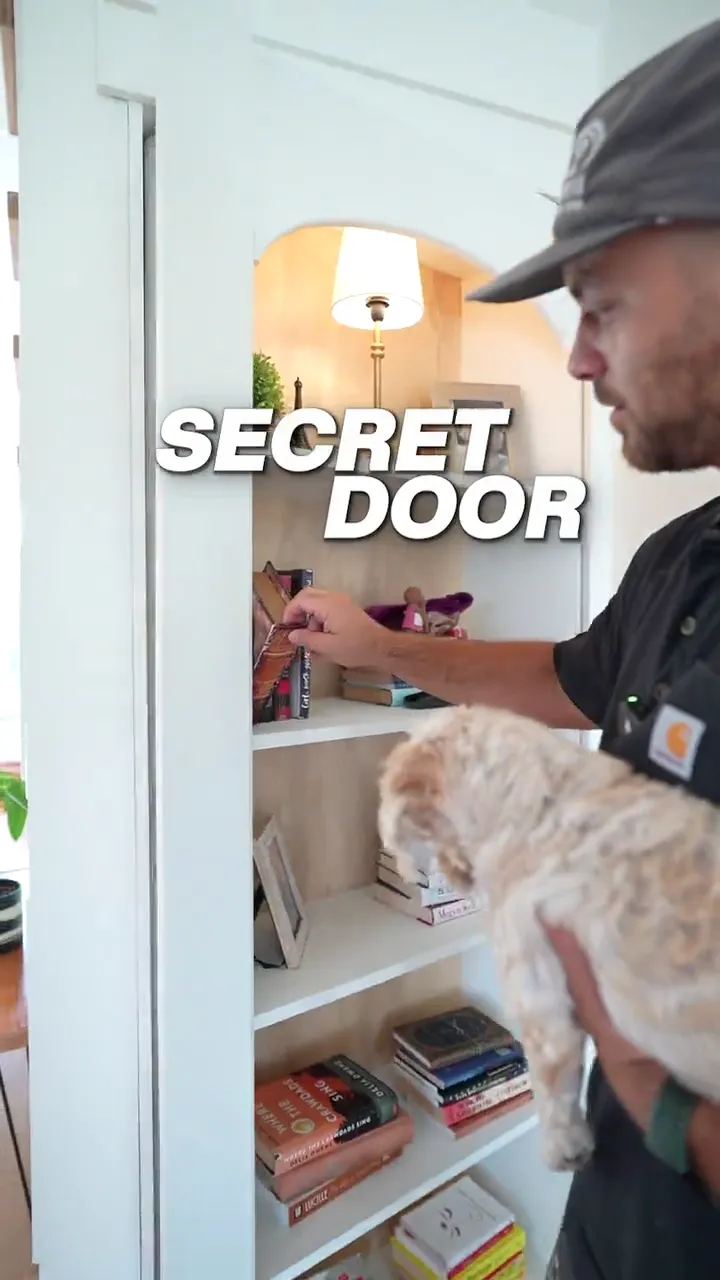 Benny of Jetty Woodwork opening the hidden bookcase door he built under a staircase, holding a small dog